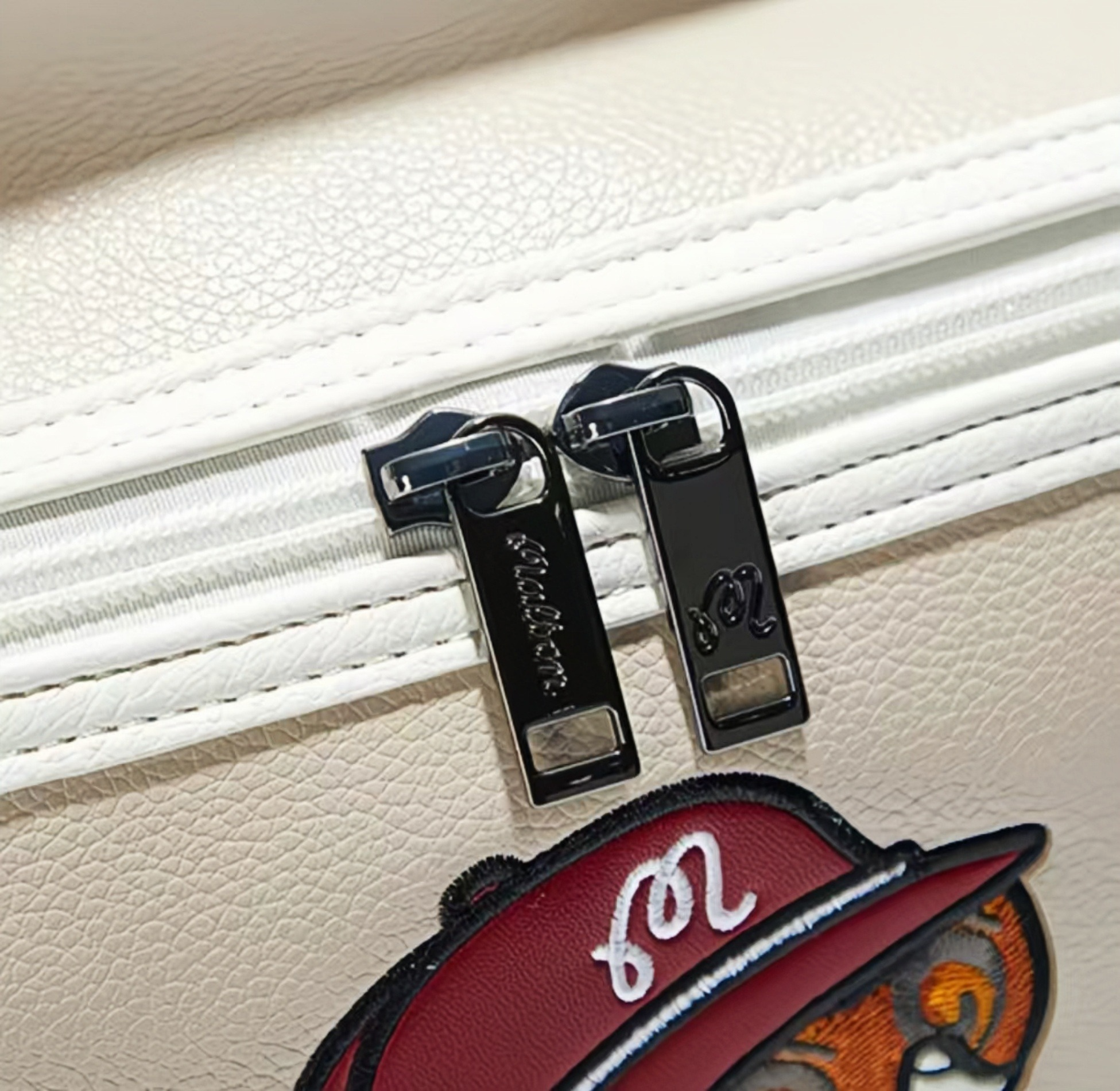 Detail shot of Malbon Golf light pouch showing dual black zipper pulls with engraved "Malbon" logo, white stitching, and premium PU leather texture