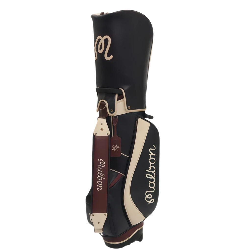 Front view of Malbon Golf stand bag in black PU leather with cream trim, showing the "Malbon" script logo, brown shoulder strap, and vintage-style tag with brand emblem.