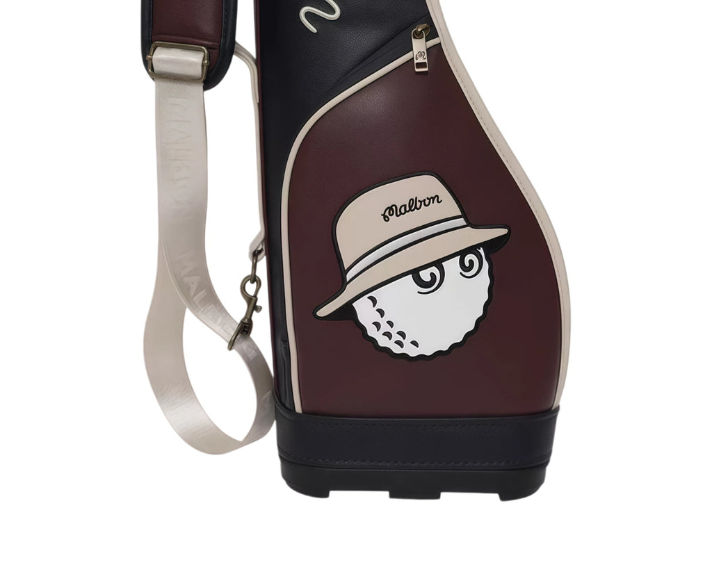 Front close-up of Malbon Golf Classic Sunday bag showcasing embroidered golf ball mascot wearing a beige hat with "Malbon" text. Fun and fashionable detail for golf lovers.