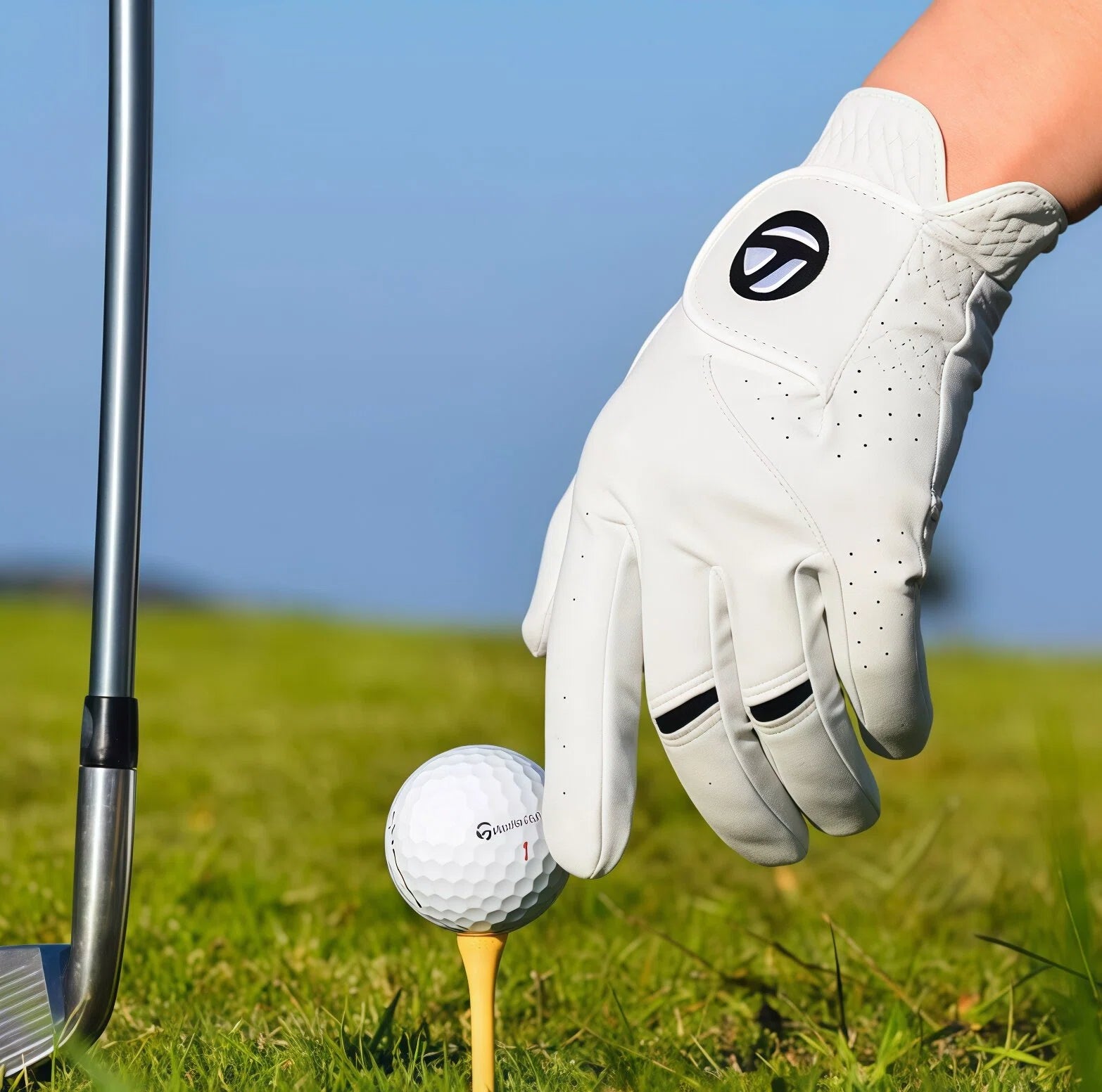 Golf glove, golf ball, and club on a golf course with a clear blue sky.