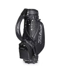 Full view of the Titleist Heritage Cart Bag in black glossy finish. Features the "Titleist" script logo in silver, padded shoulder strap with "Vokey Design" text, and multiple zippered compartments. Built for durability and elegance.